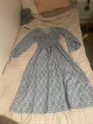 Blåvit långklänning med mönster - Supersöt långklänning i blått och vitt med ett snyggt grafiskt mönster. Klänningen har lång ärm, smockad midja och v-ringning med knappar framtill. Perfekt för dig som gillar boho-vibes och vill ha något luftigt och bekvämt.