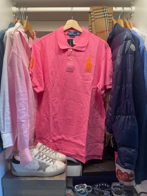 Rosa pikétröja från Polo Ralph Lauren - Snygg rosa pikétröja från Polo Ralph Lauren i storlek XXL. Med klassisk krage och knappar framtill. Stor orange broderad polospelare på bröstet och gul broderad siffra på ärmen. Perfekt till sommaren och på en vanlig vardag. Nypris ligger runt 1400. Tröjan är aldrig använd och har pristagarna kvar.                                                           Skriv vid frågor, priset är inte hugget i sten🤝