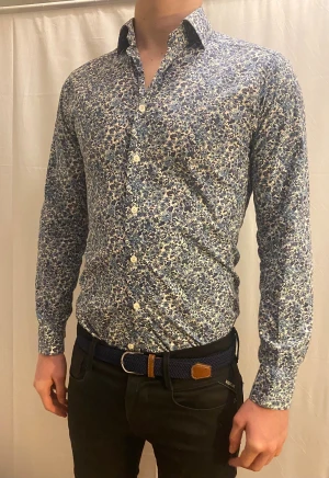 Blommig slim fit skjorta från Eton - Snygg skjorta från Eton med blommigt mönster i blå och lila toner på vit botten. Slim fit-modell med klassisk krage och knappar framtill. Tillverkad i mjuk bomull som känns skön mot huden. Perfekt för dig som gillar detaljer och vill sticka ut lite extra. Pris kan diskuteras vid snabb och smidig affär. Perfekt julklapp 🎄🎁