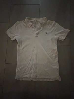 Vit piké med liten broderad logga. Pojke - Vit pikétröja med klassisk krage och korta ärmar. Framtill finns en liten broderad logga i svart på vänster bröst. Tröjan har knappar vid halsen och är gjord i mjukt bomullsmaterial. Perfekt för en clean och enkel stil.