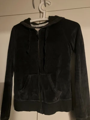 Svart velour hoodie från Hollister - Svart hoodie från Hollister i mjuk velour med dragkedja framtill, huva med snörning och två fickor. Tröjan har ribbade muddar och en liten metall-logga vid fickan. Perfekt för chill dagar och sköna outfits.