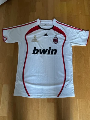 AC Milan vit Adidas - AC Milan bortatröja i vitt med tunna vertikala ränder, röda och svarta detaljer samt klubbmärke och Champions League-brodyr. Adidas-logga på bröstet och 'bwin' som sponsor. Tillverkad i lätt och ventilerande polyester, kortärmad modell.