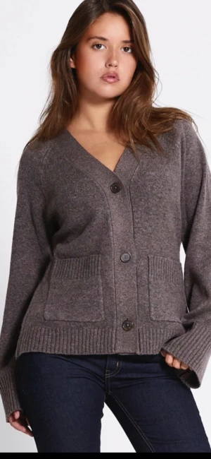 Brun kofta med knappar och fickor - Mysig brun kofta med v-ringning, stora knappar framtill och två rejäla fickor. Koftan har ribbade muddar och är långärmad, perfekt att slänga på sig när det är kyligt. Materialet känns stickat och ger en avslappnad vibe.