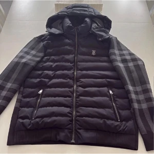 Svart pufferjacka från Burberry - Säljer en svart pufferjacka från Burberry med ikoniskt rutigt mönster på ärmar och rygg. Jackan har huva, två dragkedjeförsedda fickor och silverfärgad TB-logga på bröstet. Priset kan diskuteras 