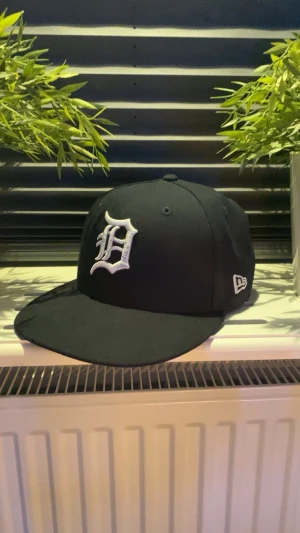 Blå New Era 59FIFTY Detroit keps - Snygg marinblå New Era 59FIFTY keps med Detroit Tigers broderad D-logga i vitt framtill och MLB-logga baktill. Klassisk flat brim och strukturerad form. Tillverkad i polyester, perfekt för dig som gillar streetwear och sportig stil.          Storlek 7 och kepsen är inte använd mycket. Hör av dig om du har några frågor så svarar jag snabbt. Priset är prutbart vid enkel affär