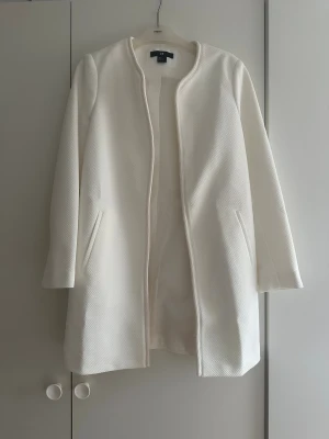 Vit kappa från H&M i strukturerad tyg - Säljer en stilren vit kappa från H&M med rak passform och diskret struktur i tyget. Jackan har lång ärm, två fickor fram och är helt utan knappar eller dragkedja för en clean look