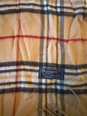 Rutig beige halsduk från Burberrys - Klassisk halsduk från Burberrys i beige med det ikoniska rutmönstret i svart, vitt och rött. Gjord av 100% lammull, mjuk och varm med fransar i ändarna. Perfekt för dig som vill ha en tidlös och snygg accessoar med brittisk vibe.