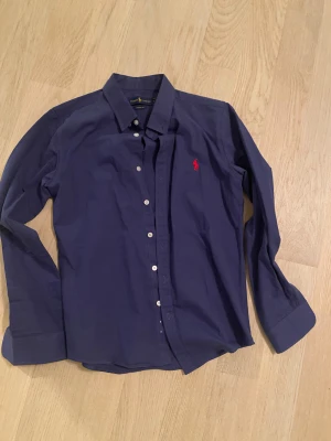 Blå skjorta från Ralph Lauren M - Snygg blå skjorta från Ralph Lauren i klassisk modell med vit knappstängning och röd broderad logga på bröstet. Skjortan har lång ärm, krage och är gjord i mjuk bomull. Perfekt för dig som gillar stilrena och tidlösa plagg.