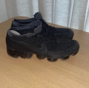 Nike Vapormax svarta sneakers - Svarta Nike Vapormax sneakers med stickad ovandel och synlig bubblig sula. Snyggt diskret Nike-logga på sidan och Vapormax-tryck på hälen. Skorna har snörning och rund tå, och är tillverkade i syntet och stickat material för en modern look. Tveka inte på att höra av dig!