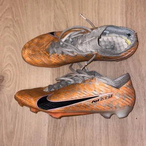 Nike Air Zoom orange fotbollsskor - Säljer ett par Nike Air Zoom fotbollsskor i orange och grått med svart swoosh. Skorna har snörning, platt sula med dobbar och ett mönstrat syntetmaterial. Perfekt för dig som vill sticka ut på planen med snygg design och bra grepp.