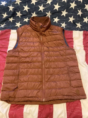 Brun quiltad dunväst Dressmann XL - Quiltad brun dunväst från Dressmann i storlek XL. Utsidan är brun med horisontella sömmar och dragkedja framtill, insidan är mörkblå. Hög krage med ribbad insida för extra komfort. Perfekt för lager-på-lager under kyliga dagar. Västen är i storlek XL och är väldigt väl behandlad. 