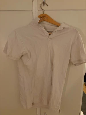 Vit kortärmad  skjorta - Vit  skjorta med korta ärmar och klassisk krage. Skjortan har tre knappar vid halsen och är gjord i mjukt bomullstyg. Passformen är normal och enkel att matcha med jeans eller shorts. Perfekt för dig som gillar stilrena och enkla plagg.