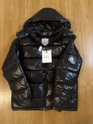 Moncler Maya Jacka - Svart pufferjacka från Moncler med glansig finish, huva och dragkedja framtill. Jackan har Moncler-logga på ärmen och praktisk ficka med lock. Perfekt för kalla dagar och har en riktigt schysst streetstil.