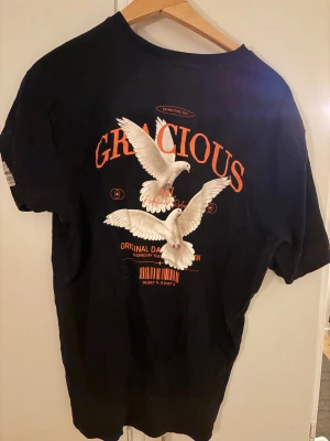 Svart t-shirt med tryck från Gracious - Svart t-shirt från Gracious med coolt tryck av två vita duvor och orange text på ryggen. Framtill finns en mindre logga och textdetaljer. T-shirten har korta ärmar och är gjord i mjuk bomull, perfekt för dig som gillar streetwear med statement-prints.