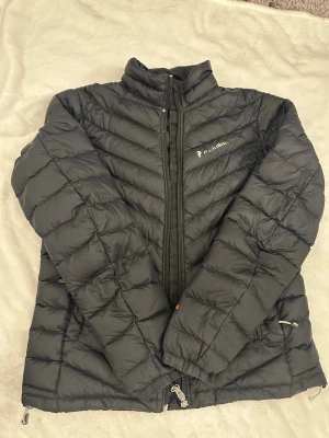Svart dunjacka Peak Performance S - Svart dunjacka från Peak Performance i storlek S. Jackan har quiltad design med dragkedja framtill och hög krage. Perfekt för kalla dagar och har en snygg, sportig look. Materialet är lätt och håller värmen bra. Passar dig som gillar stilren och funktionell ytterjacka.