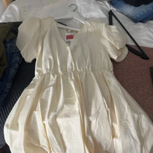 Ljus kortklänning med puffärm - Supersöt ljus kortklänning från H&M med puffiga kortärmar och v-ringad hals. Klänningen har en markerad midja och är gjord i ett lätt, glansigt material som ger en fräsch och somrig vibe. Perfekt för dig som gillar enkel men trendig stil.