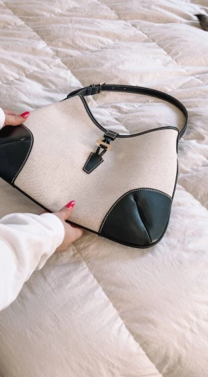 Beige och svart axelväska med skinn - Stilren axelväska i beige canvas med svarta skinn-detaljer och guldfärgat spänne framtill. Väskan har en justerbar axelrem och snygga kontrastsömmar. Väskan är från H&M, ordinarie pris 349, jag säljer den för 100 kr, pris går att diskutera. Använt endast 2 gånger. 