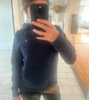 Mörkblå hoodie från Gant - Säljer en mörkblå hoodie från Gant med klassisk logga på bröstet. Tröjan har huva, långärmad design och en stor magficka framtill. Perfekt för chill dagar och enkel att matcha med jeans eller joggers.