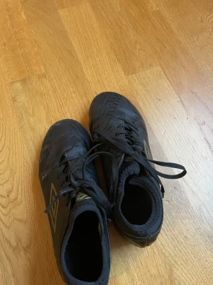 Svarta fotbollsskor Umbro strl 35 - Umbro Raposa SC AG fotbollsskor i svart med gulddetaljer och snörning. Skorna har rund tå och platt sula med fasta dobbar för bra grepp på konstgräs. Tillverkade i syntetmaterial med mönstrad ovandel och Umbro-logga på sidan. Perfekt för dig som vill ha stil på planen.