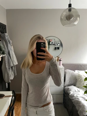 Ljusgrå/beige långärmad topp med rund hals - En basic ljusgrå långärmad topp med rundad, djup urringning. Tunn och mjuk i materialet, perfekt att styla med jeans eller kjol. Sitter figurnära och har en enkel, stilren look utan synliga detaljer eller mönster.