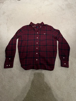 Röd rutig skjorta från GANT i stl 170 (15år) - Snygg röd och mörkblå rutig skjorta från GANT med klassisk button-down krage och vita knappar. Skjortan har lång ärm och broderad GANT-logga på bröstet. Tillverkad i mjuk bomull som är skön mot huden. Perfekt för dig som gillar stilrena och tidlösa plagg. Knappt använd