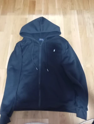 Svart hoodie från Polo Ralph Lauren - Svart hoodie från Polo Ralph Lauren med dragkedja framtill och klassisk huva med snörning. Tröjan har en vit broderad logga på bröstet och praktisk känguruficka. Perfekt för chill dagar och enkel att matcha med jeans eller joggers.