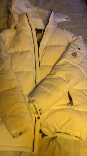 Vit Moncler pufferjacka med huva - Säljer en riktigt snygg vit pufferjacka från Moncler. Jackan har en stor huva, svarta detaljer på dragkedjorna och Moncler-logga på ärmen. Insidan är svart med en cool serieteckning. Perfekt för kalla dagar när du vill hålla stilen.
