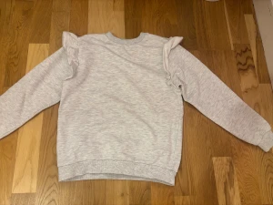 Ljusgrå volangtröja från H&M - Superfin ljusgrå tröja från H&M i mjuk ekologisk bomull. Tröjan har långa ärmar och snygga volangdetaljer på axlarna som ger en extra touch. Perfekt för dig som gillar en enkel men ändå söt stil.