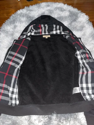 Svart rutig hoodie från Burberry - Svart hoodie från Burberry med klassiskt rutigt foder i svart, vitt och rött. Jackan har huva, dragkedja och fickor framtill. Materialet är 100% bomull och insidan är mjuk. Perfekt för dig som gillar stilren streetwear med lyxig touch.
