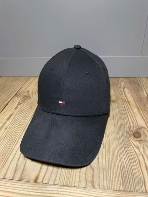 Svart keps Tommy Hilfiger - Svart keps från Tommy Hilfiger med klassisk böjd skärm och broderad logga i vitt, rött och blått framtill. Tillverkad i bomull med snygga sömmar och stilren design. Perfekt för dig som vill ha en clean och enkel look.