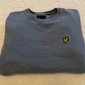 Blå sweatshirt från Lyle & Scott - Säljer en stilren blå sweatshirt från Lyle & Scott med klassisk rund halsringning och broderad gul logga på bröstet. Tröjan har ribbade muddar och är tillverkad i mjuk bomull, perfekt för en avslappnad look.