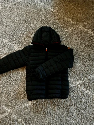 Save The Duck jacka - Svart pufferjacka från Save The Duck med huva och orange logopatch på ärmen. Jackan har quiltad design, hel dragkedja framtill och är tillverkad i syntetmaterial. Perfekt för kalla dagar. Är i barn storlek men skulle säga att den passar i xs. Hör av er vid frågor ✌️