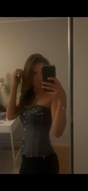 Blå denim tubtopp med knappar - Säljer en snygg blå tubtopp i denim med silverfärgade knappar framtill och rå kant nedtill. Toppen har en cool korsettliknande passform och är axelbandslös, perfekt för dig som gillar statement-plagg. Den har även en liten fickdetalj upptill för extra stilpoäng.