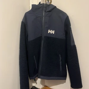 Mötkblå fleecejacka från Helly Hansen - Säljer en mörkblå fleecejacka från Helly Hansen med dragkedja och huva. Jackan har långa ärmar och är perfekt för kyligare dagar. Den har en stilren design med Helly Hansen-logga på bröstet.
