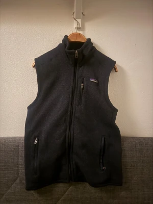 Svart fleeceväst från Patagonia  - Svart fleeceväst från Patagonia i storlek S. Västen har hög krage, hel dragkedja framtill och tre praktiska fickor med dragkedja. Klassisk Patagonia-logga på bröstet. Perfekt för lager-på-lager och outdoor-vibes.