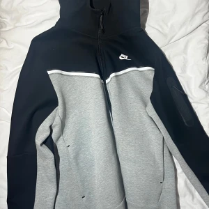 Nike hoodie med dragkedja, svart/grå - Nike hoodie med huva och hel dragkedja, svart upptill och grå nedtill med vit detalj över bröstet. Snygg logga på bröstet och praktisk ficka med dragkedja på ärmen. Perfekt för chill eller träning.