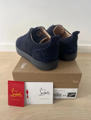 Mörkblå sneakers från Christian Louboutin - Snygga mörkblå sneakers från Christian Louboutin i mocka med klassisk röd sula och broderad logga på hälen. Skorna har rund tå, snörning och platt sula. Perfekt för dig som vill sticka ut med exklusiv design och lyxiga detaljer.