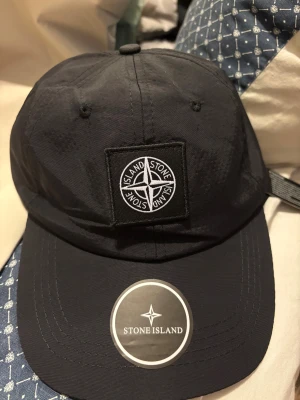 Svart keps från Stone Island - Svart keps från Stone Island med klassisk broderad logotyp framtill. Justerbar rem baktill för perfekt passform. Kepsen är tillverkad i lätt och slitstark polyester, perfekt för en clean och sportig look. Snygga sömmar och detaljer gör den till ett måste i garderoben.