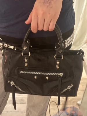 Svart handväska med dragkedjor - Cool svart handväska i skinnimitation med silvriga nitar och flera dragkedjor framtill. Väskan har två rejäla handtag och en rymlig insida med innerfack. Perfekt för dig som gillar edgy detaljer och vill ha plats för allt viktigt. Pris kan diskuteras!!💕