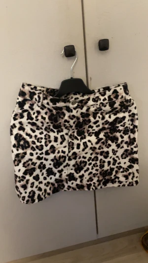 Leopardmönstrad kortkjol i bomull - Snygg kortkjol med leopardmönster i svart, beige och bruna toner. Kjolen har klassisk jeansmodell med knapp och dragkedja framtill samt fickor. Perfekt för dig som vill sticka ut med en trendig look.