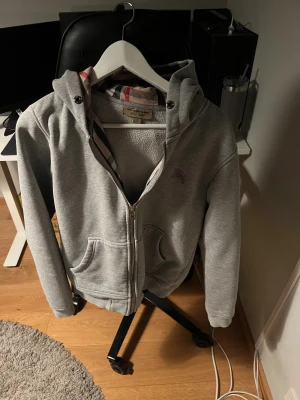 Grå hoodie från Blurberry med dragkedja - Säljer en grå hoodie från Blurberry med klassiskt rutigt foder i huvan. Tröjan har dragkedja framtill, två fickor och ribbade muddar. Perfekt för dig som gillar stilrena och lyxiga streetwear-plagg. Ikonisk Blurberry-logga broderad på bröstet.