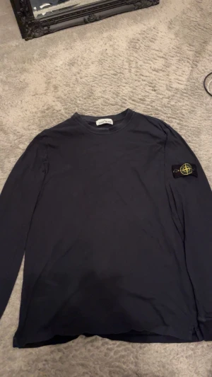 Stilren Stone Island  - Svart långärmad tröja från Stone Island i storlek XL. Tröjan har klassisk rund hals och den ikoniska Stone Island-patchen på vänster ärm. Tillverkad i mjuk bomull för skön känsla och clean look. Perfekt för dig som gillar streetwear och stilrena plagg.