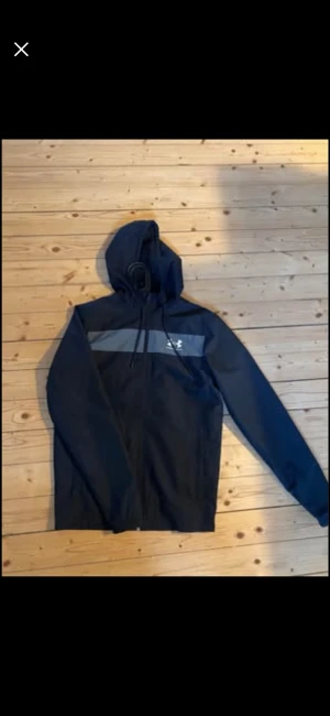 Svart vindjacka från Under Armour - Säljer en svart vindjacka från Under Armour med huva och dragkedja framtill. Jackan har en grå horisontell detalj över bröstet och logga på vänster sida. Perfekt för dig som vill ha en sportig och clean look. Långärmad och tillverkad i lätt polyester. Endast provad 1 gång och tar gärna emot bud, det är bara att skriva. Vid fler bilder skriv gärna