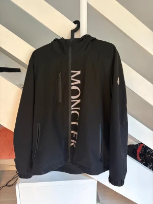 Svart vindjacka från Moncler med huva - Säljer en svart vindjacka från Moncler med stor logga framtill och broderad patch på ärmen. Jackan har huva, dragkedjor på fickorna och bröstficka samt justerbara muddar. Perfekt för dig som vill ha en clean och stilren look med exklusiv känsla.