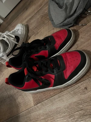 Nike sneakers röd och svart - Snygga Nike sneakers i rött och svart med klassisk swoosh på sidan. Skorna har snörning, rund tå och platt sula. Ovandelen är i syntet och skinn, med perforeringar på tån för extra ventilation. Perfekt för dig som gillar sportig och cool stil.