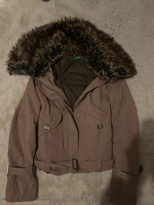 Brun parkajacka med fuskpäls från Benetton - Parkajacka i brun färg från United Colors of Benetton med stor huva kantad av fuskpäls. Jackan har bälte i midjan, knappar på fickorna och detaljer vid ärmsluten. Materialet är bomull och syntet, perfekt för kyliga dagar.