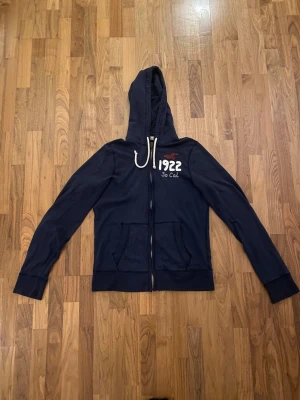 Marinblå hoodie från Hollister - Säljer en marinblå hoodie från Hollister med broderad logga och text '1922 So Cal' på bröstet. Tröjan har huva med snörning, två fickor fram och dragkedja. Materialet är mjuk bomull och modellen är normal passform med långa ärmar.