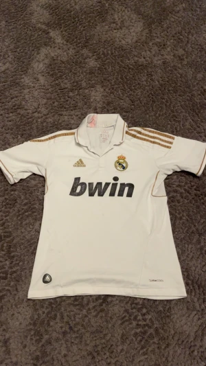 Real Madrid Ronaldo 7 Adidas tröja - Säljer en vit Real Madrid fotbollströja från Adidas med guldiga detaljer och tre ränder på axlarna. Tryck med 'bwin' på bröstet, klubbmärke och LFP-patch på ärmen. På ryggen står det 'Ronaldo' och nummer 7 i guld. Tillverkad i lätt ClimaCool-material.