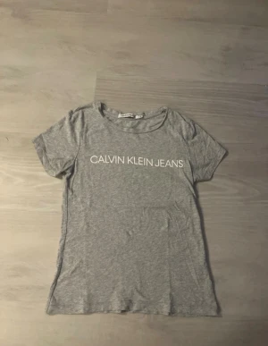Grå t-shirt Calvin Klein Jeans XS - Snygg grå t-shirt från Calvin Klein Jeans i storlek XS. T-shirten har korta ärmar och rund hals, med vit logotyp framtill. Tillverkad i mjuk bomull som känns skön mot huden. Perfekt för en chill och stilren look.