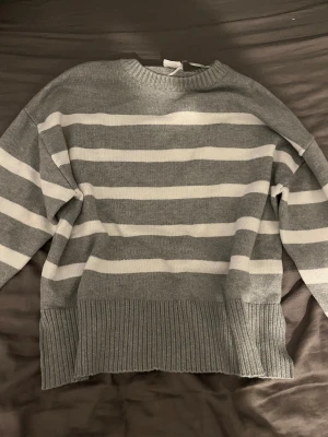 Gråvit randig stickad tröja Stories S - Supermysig stickad tröja från Stories i storlek S. Tröjan är grå med breda vita ränder, har rund hals och långa ärmar. Ribbad kant vid hals, ärmslut och nederkant ger en snygg touch. Perfekt för dig som gillar chill och stilren vibe.